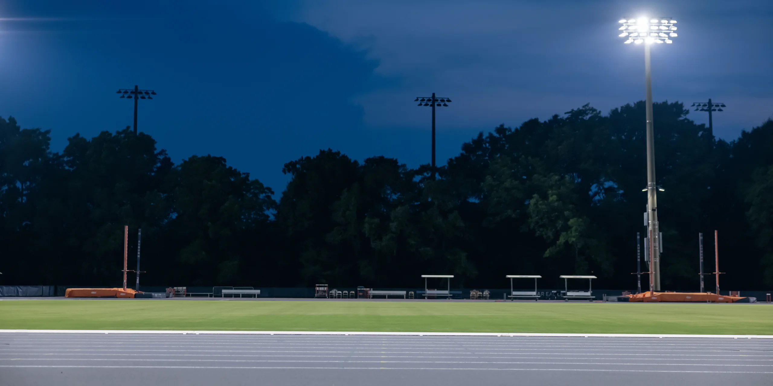 A sprinter in an orange Outperform singlet, shown at seven points of one flying sprint under the floodlights